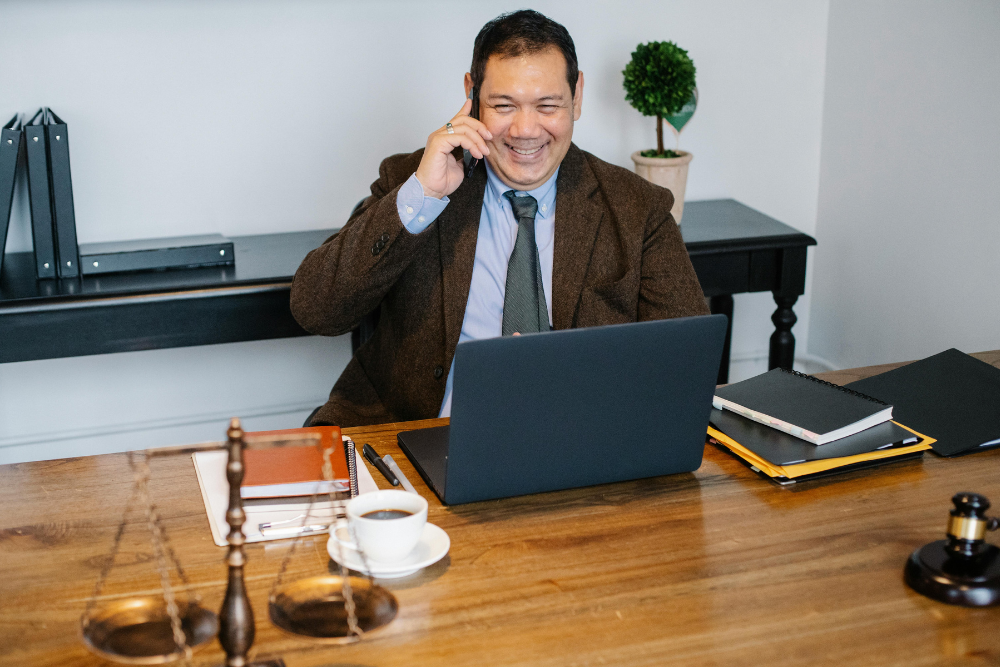 A smiling male lawyer in a brown blazer talking on a smartphone while sitting at a desk