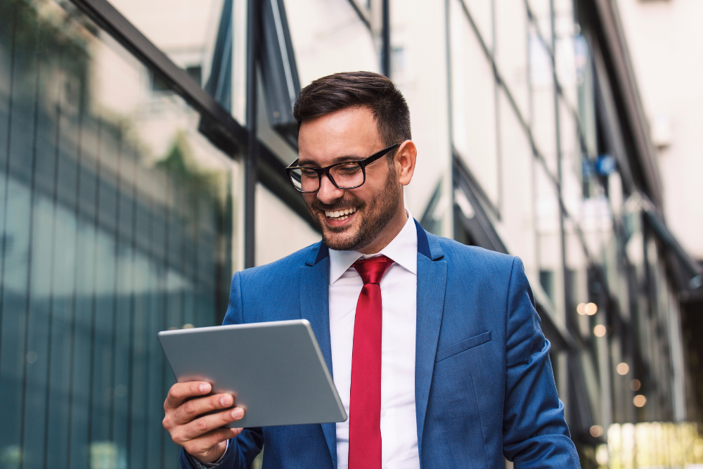 A smiling man in a blue suit, white shirt, and red tie holding a tablet while standing in front of a modern glass building.