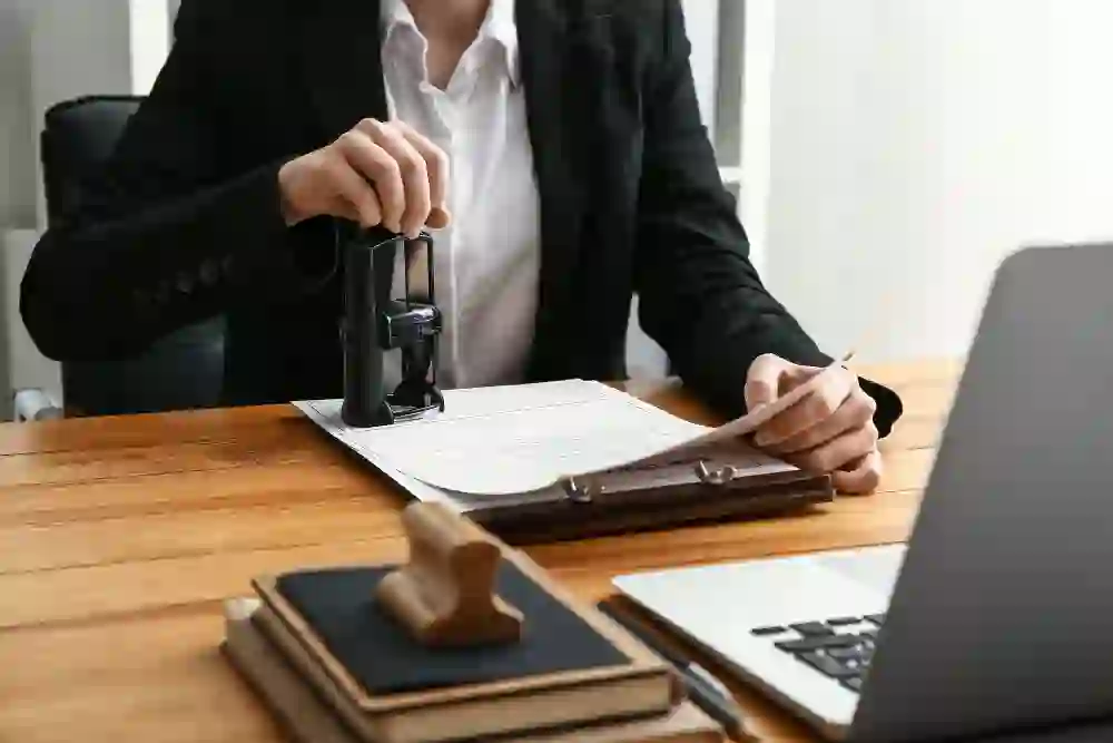 professional in a black blazer using a large self-inking stamp to certify a legal document on a clipboard at a wooden desk