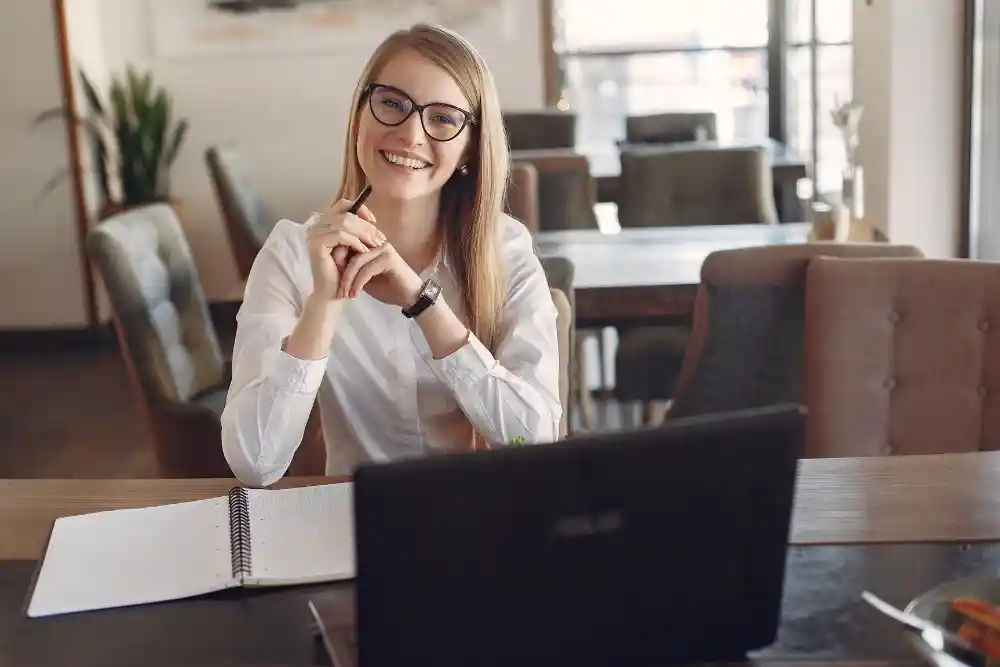 A smiling young professional woman with glasses sitting at a table with a laptop and notebook
