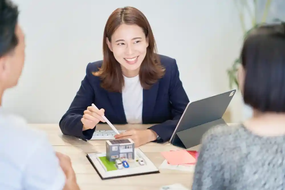A smiling real estate agent or architect in a navy blazer presenting a miniature house model and digital tablet to a couple during a consultation.