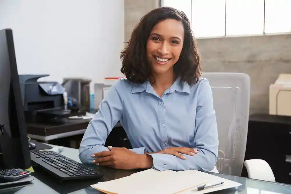 A smiling professional woman in a light blue button-down shirt sitting at an office